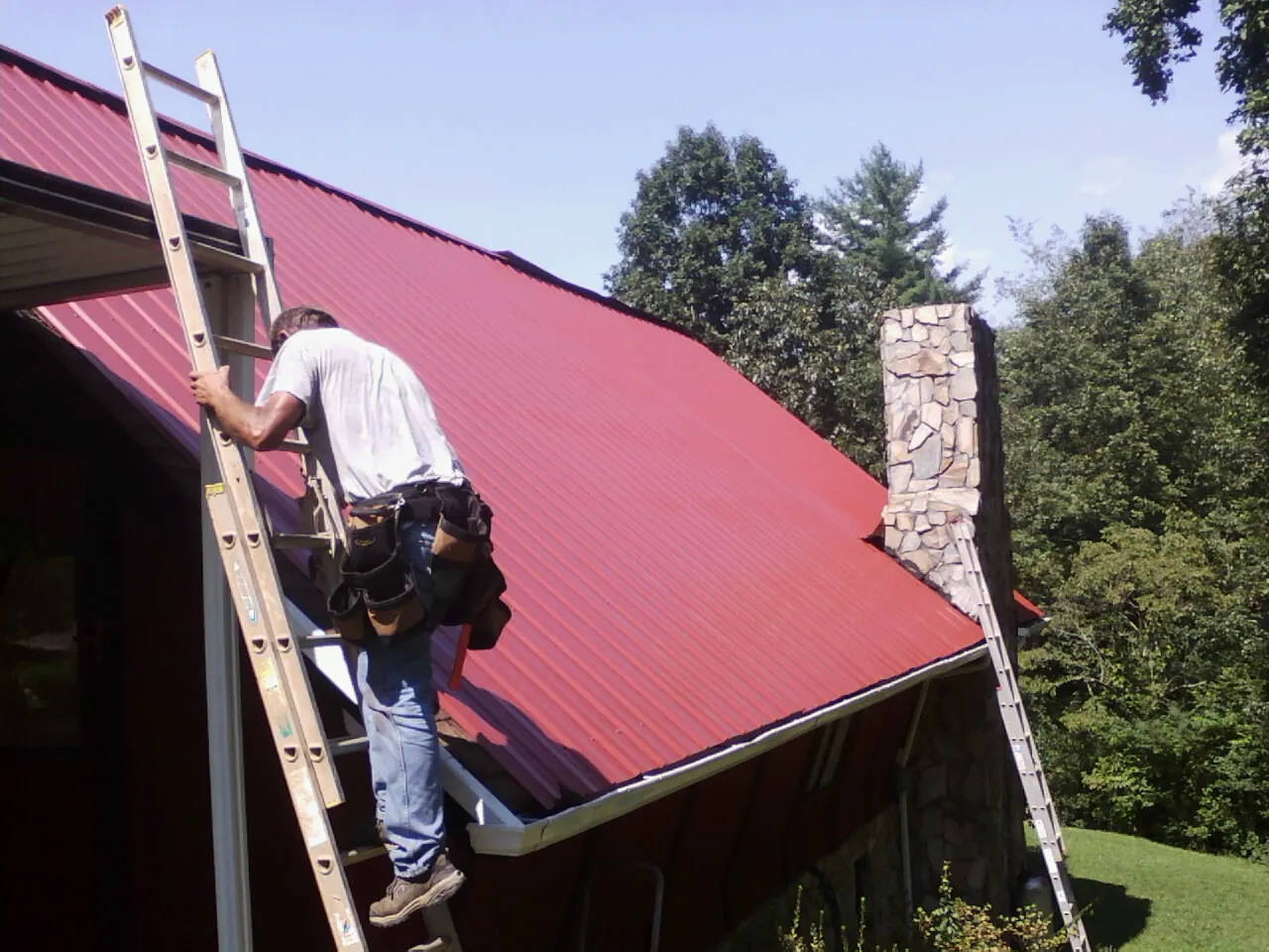 Roofer on a ladder beside a red metal roof and stone chimney — Soffit & Fascia Repair by Roofing Brothers in Crozier
